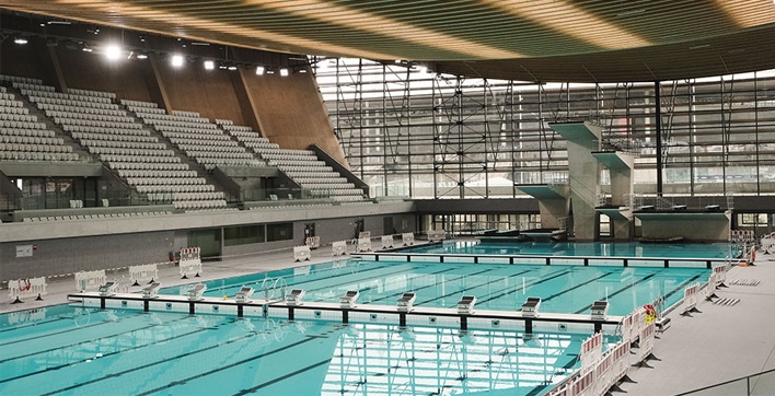 Photo du Centre Aquatique Olympique de Saint-Denis, une piscine moderne et spacieuse avec un grand bassin et un plongeoir à plusieurs hauteurs, baignée de lumière. Elle est reliée au Stade de France par une passerelle visible en arrière-plan.