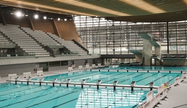Photo du Centre Aquatique Olympique de Saint-Denis, une piscine moderne et spacieuse avec un grand bassin et un plongeoir à plusieurs hauteurs, baignée de lumière. Elle est reliée au Stade de France par une passerelle visible en arrière-plan.