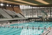 Photo du Centre Aquatique Olympique de Saint-Denis, une piscine moderne et spacieuse avec un grand bassin et un plongeoir à plusieurs hauteurs, baignée de lumière. Elle est reliée au Stade de France par une passerelle visible en arrière-plan.